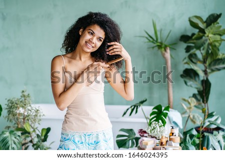 Haircare, morning routine, eco friendly zero waste concept. American multiethnic female combing brown thick curly hair with wooden comb on green natural bathroom interior with plants on background.