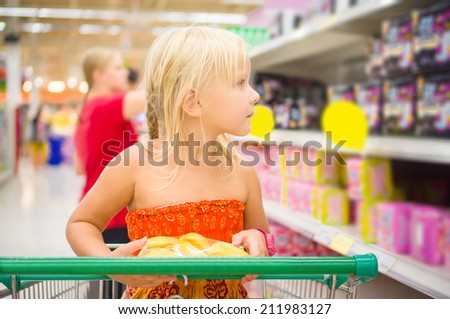 Adorable girl in shopping cart looks at goods on shelves in supermarket