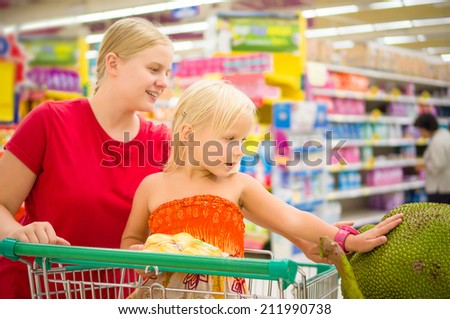 Young mother and adorable girl in shopping cart looks at giant jack fruits on boxes in supermarket