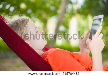 Young woman reading on electronic tablet reader relaxing in hammock under palm trees
