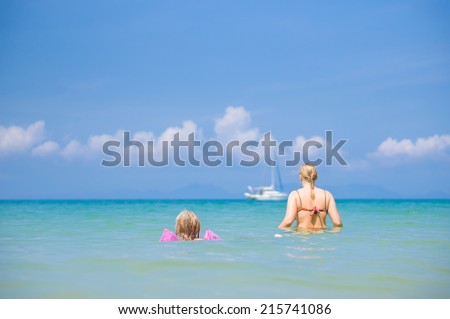 Young mother and daughter swim in clear tranquil ocean with yacht on back