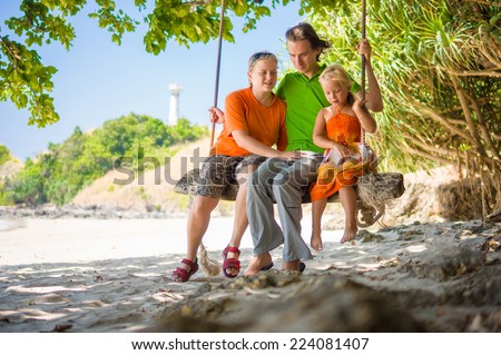 Mother, father and daughter embraces on rope swing under palm trees on tropical beach with lighthouse on back