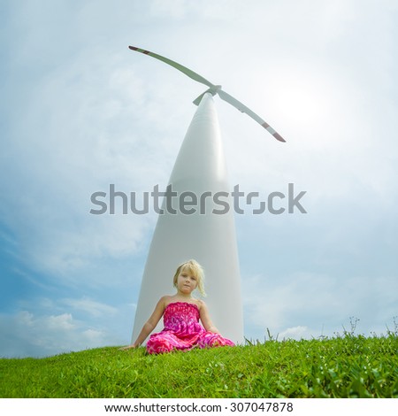 Adorable girl sit on grass with giant white wind turbine generating electricity on back