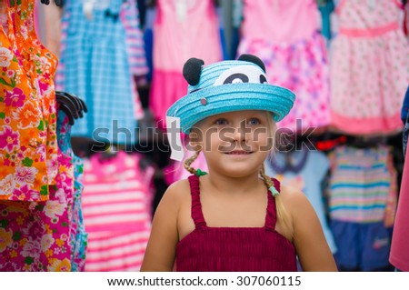 Adorable girl selects blue hat among beautiful baby girl dresses on stands and hangers in supermarket