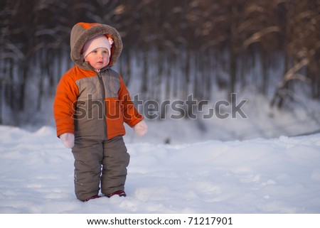 Cute adorable baby stay near ski protection fence with forest on background and look to skiers on mountain track