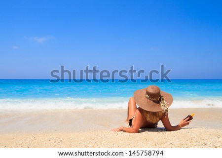 Woman laying on the beach in beach brown straw hat holding a sun lotion bottle enjoying summer holidays looking at the ocean