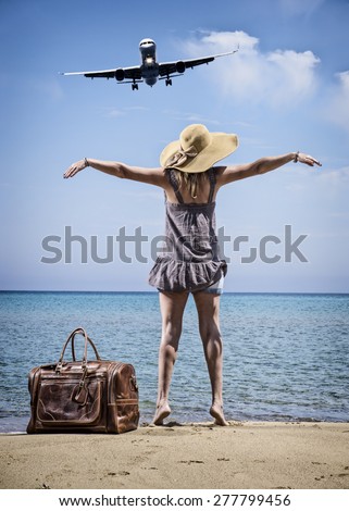 Woman with vintage leather travel bag on the beach looking at an airplane passing by