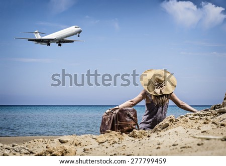 Woman with vintage leather travel bag on the beach looking at an airplane passing by
