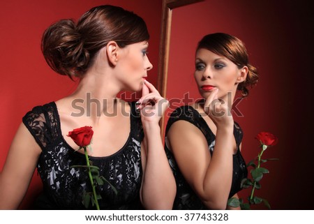 A beautiful sexy women wearing an evening dress and holding a red rose looking at her reflection in the miror on red background