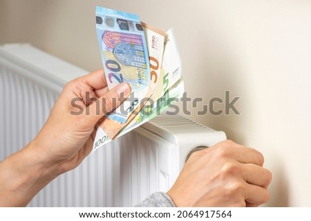 Woman hand holding euro banknotes and adjusting temperature of central heating radiator at home. Family pay money for home heating. Expensive heating costs 商業照片 © 