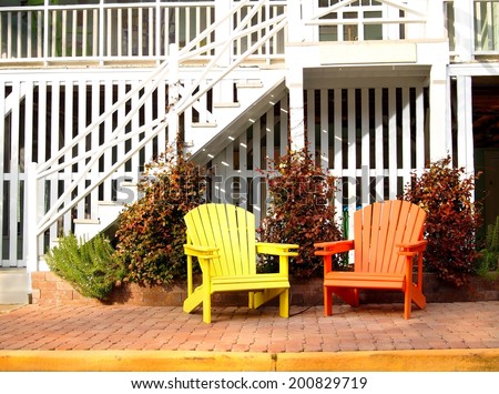 A pair of wooden adirondack chairs, one yellow and one orange, in front of a white beach house.