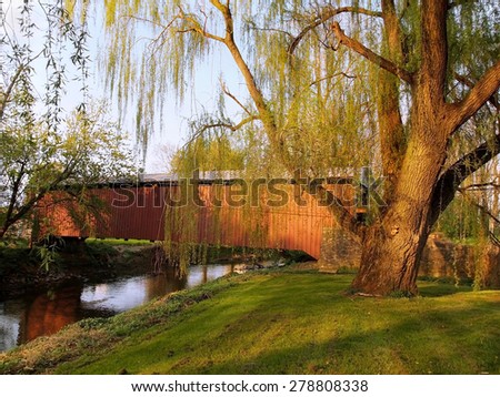 A beautiful willow tree with a swing stands beside the Lime Valley Covered Bridge, also known as Strasburg Bridge, in Lancaster County, Pennsylvania.