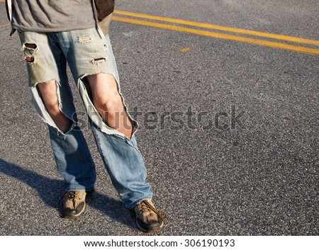 A young man, from the waist down,  in a causal t-shirt and ripped jeans with hair legs exposed stands in the road.