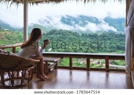 Portrait image of a beautiful asian woman holding and drinking hot coffee , sitting on balcony and looking at mountains and green nature 