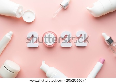 Best skincare products and cosmetic trends of 2022 concept. 2022 white number with lip balm, cream bottle, serum and lotion on pink background.