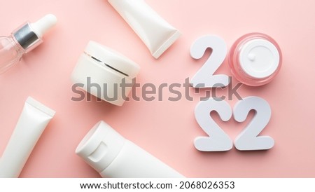 Best skincare products and cosmetic trends of 2022 concept. 2022 white number with lip balm, cream bottle, serum and lotion on pink background. Love your skin.