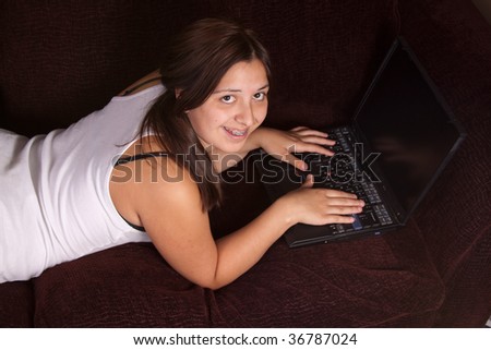 Pretty brunette teenage girl laying on the sofa and typing on laptop