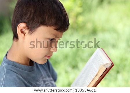 Young boy reading book outside with natural green background and shallow depth of field - with copy space to right