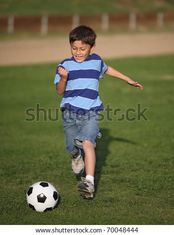 Authentic happy Latino boy playing with soccer ball in field wearing blue striped tee shirt.