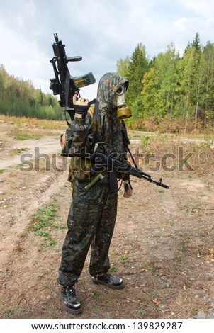 A soldier stands alone wearing state of the art bullet proof body armor, camouflage, and gas mask, holding an automatic weapon.