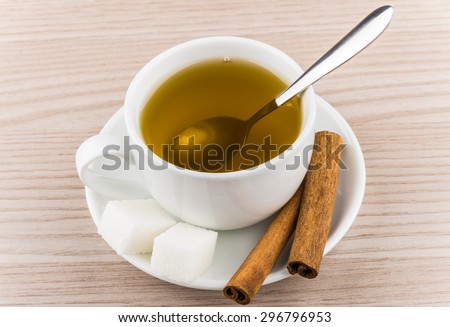 Cup of hot green tea, sugar cubes and cinnamon sticks in saucer on wooden table