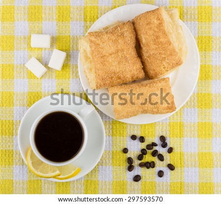Black coffee, pieces of lemon and sugar, plate with flaky biscuits on yellow tablecloth, top view