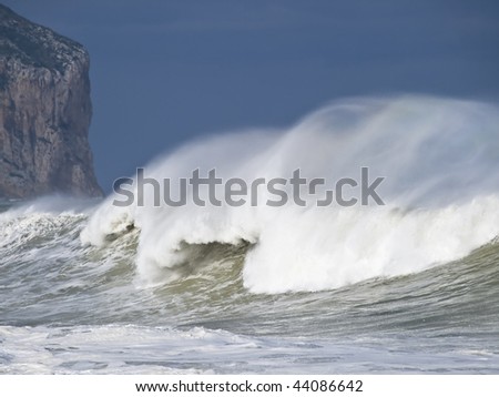 Great wave breaking at sea during a heavy storm, with wind blowing on the foam of the wave crest. In the background is the wall of a mountain cliff
