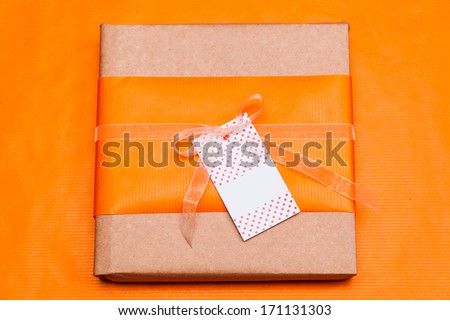 An orange paper gift box tied with a organza ribbon bow and a visiting card hooked on it in front of orange background