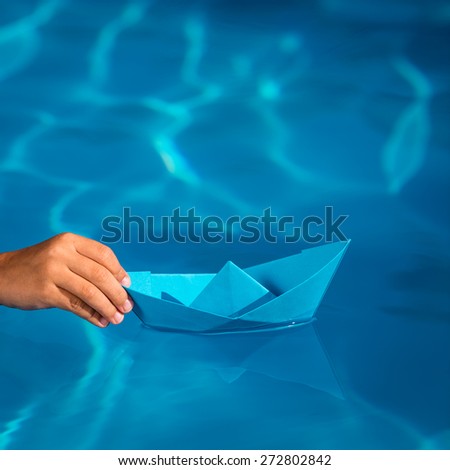 Child holding blue paper sailing boat on water background. Summer vacation and travel concept