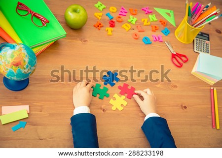 Child holding multicolor puzzles in hands. School items on wooden desk in class. Education concept. Top view