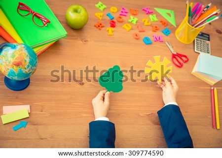 Child holding paper tree and sun in hands. School items on wooden desk in class. Education concept. Top view