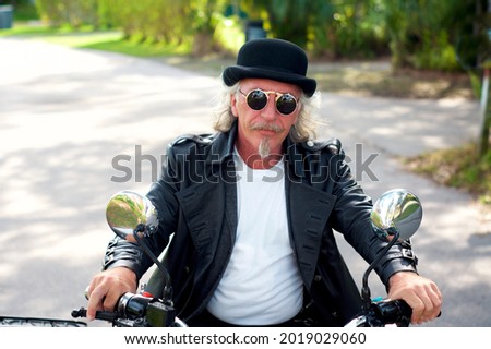 A handsome blond haired, goatee biker driving motorbike. Wearing leather tailcoat, bowler hat and round sunglasses looking at viewer with hands on handlebars. ストックフォト © 