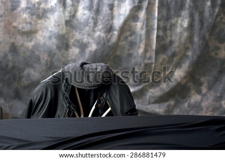 A leather motorcycle jacket hangs on the back of a chair with a tweed cap sitting on top behind a black covered table.