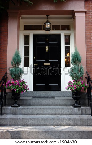Black door in Boston on a stately brownstone in the beacon hill area. Ornate flower urns and elaborate door knocker. Lights on inside the house can be seen from the street.