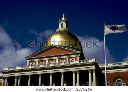 the massachusetts state house with the state flag flying to the right, the gold dome glitters in the sun in front of a deep royal blue sky with low clouds