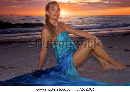 A pretty blonde woman wrapped in silk is sitting on the beach at sunset looking at the viewer with the sun visible just above the horizon.