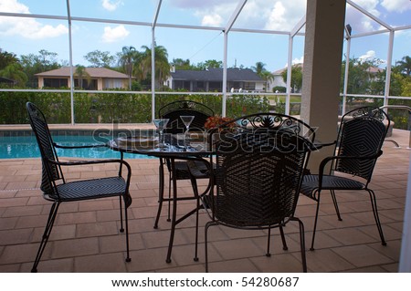 a table is set for dinner on an outside patio or lanai with swimming pool in background, the patio is screened in.