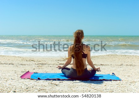 A woman with long hair is facing away from viewer looking at the sea while mediating outside at the beach in yoga lotus position.
