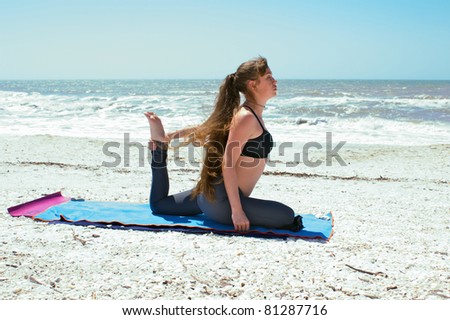 an athletic woman is doing yoga exercise on beach in Kapotasana or Pigeon Pose variation holding foot on an empty beach at the gulf of mexico in bonita springs florida with long hair blowing in wind
