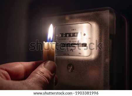 A man's hand with candle in complete darkness looking on electricity meter at home. Power outage, blackout concept. 