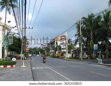 KOH SAMUI, THAILAND - OCTOBER 24, 2013: Main ring road of island with moving transport, bike, crosswalk, poles with wires on roadsides, coconut palm trees and cloudy sky in rainy season