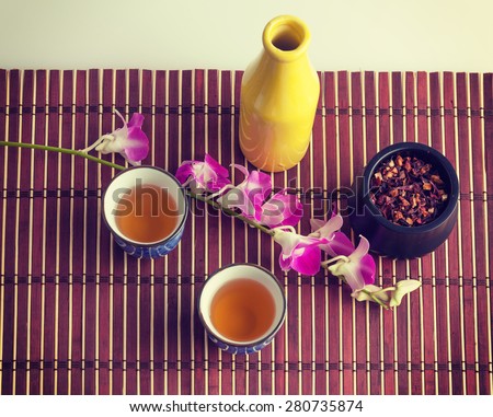 Purple orchid, yellow bottle and black tea in small Japanese cups on a bamboo mat still life composition.