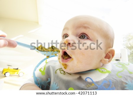 Messy baby sitting in high chair, about to eat a spoonful of food. Horizontally framed shot.