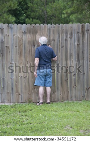 Outdoor photograph of an adult man peeking through a hole in the fence.  He is a nosy neighbor!