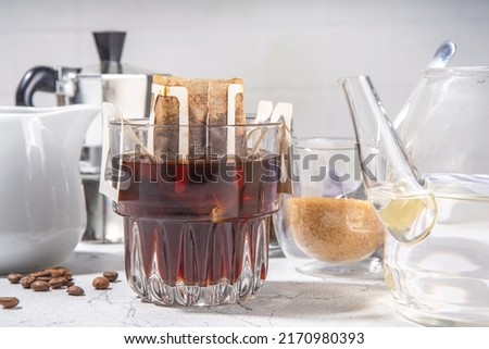 Trendy instant brewed drip coffee, cold brew coffee. Drip filter coffee bag in glass cup on white table, with brown sugar, coffee beans, pot with hot water Stock foto © 