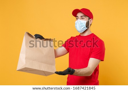 Delivery man employee in red cap t-shirt uniform mask glove hold craft paper packet with food isolated on yellow background studio Service quarantine pandemic coronavirus virus 2019-ncov concept