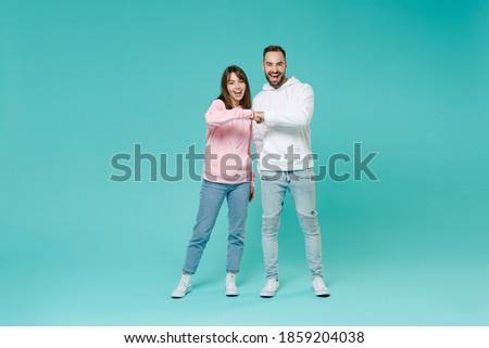 Full length of laughing funny young couple friends man woman in white pink casual hoodie holding hands folded giving fists bump looking camera isolated on blue turquoise background studio portrait