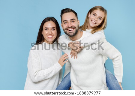 Funny young parents mom dad with child kid daughter teen girl in white sweaters giving piggyback ride to joyful, sitting on back isolated on blue background. Family day parenthood childhood concept