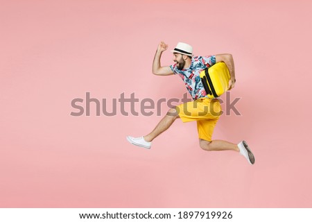 Full length side view of cheerful traveler tourist man in summer clothes hat jumping running hold suitcase isolated on pink background. Passenger traveling on weekend. Air flight journey concept