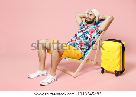 Full length of smiling young traveler tourist man in summer clothes hat sit on deck chair hold hands behind head isolated on pink background. Passenger travel on weekend. Air flight journey concept